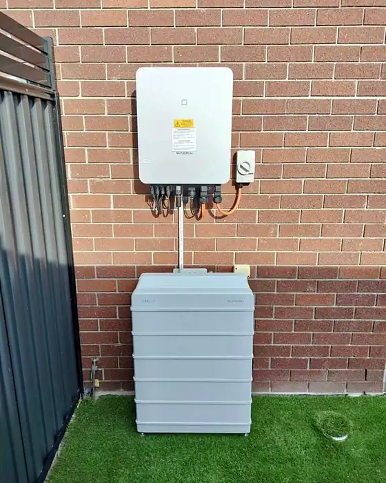 A wall-mounted hybrid inverter above a stacked home battery on a brick wall