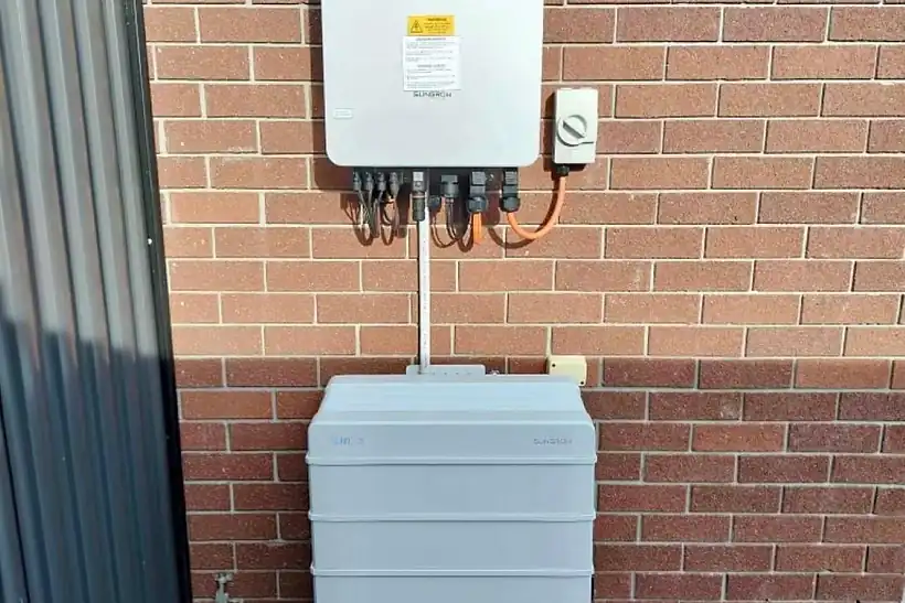 A wall-mounted hybrid inverter above a stacked home battery on a brick wall