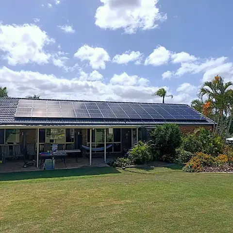 A low-set brick home with a long row of solar panels on the roof and a tropical garden in front