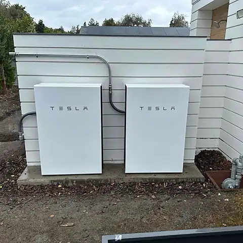 Two home batteries side by side on a slab beside a white clad house