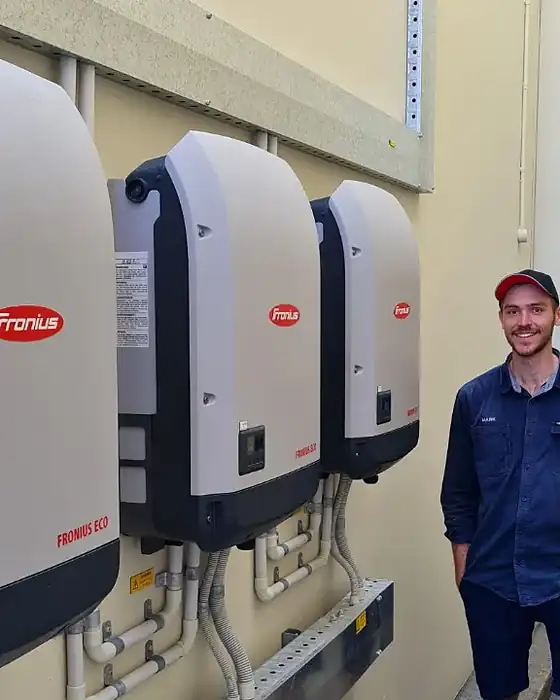 An LMS technician in a navy shirt and cap smiling beside three Fronius Eco inverters