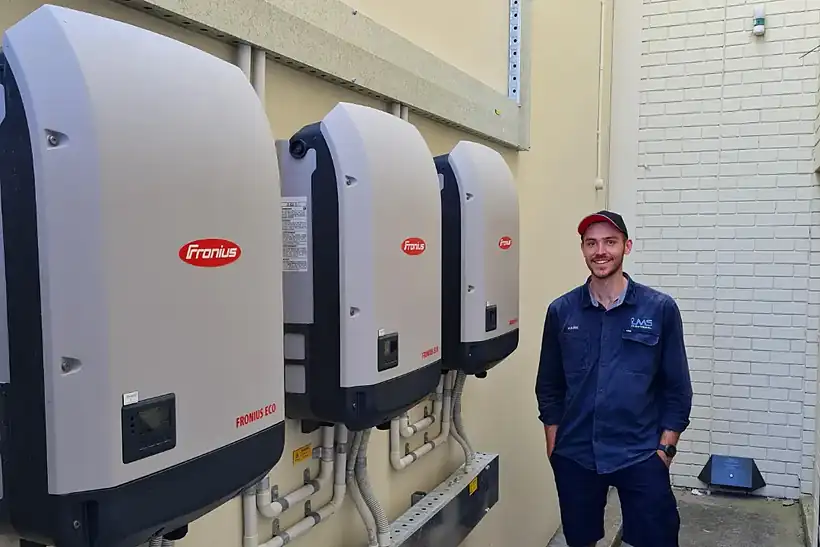 An LMS technician in a navy shirt and cap smiling beside three Fronius Eco inverters